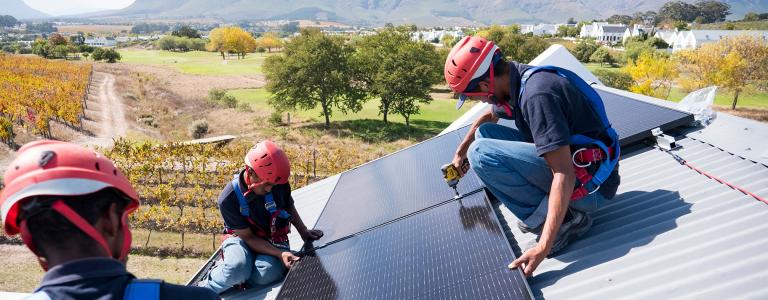 Three workers install solar panels on the roof of a house with farmland and a mountain in the background.