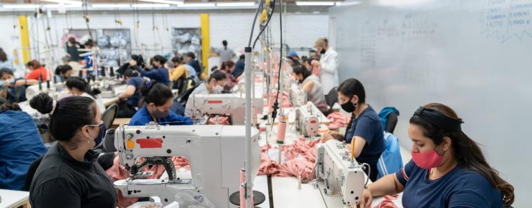 Looking down the centre of a long table lined with Latin American women working at industrial sewing machines making pink garments.