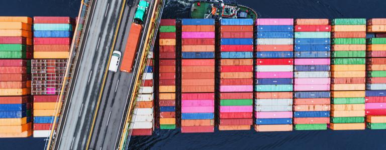 Aerial view of a cars driving across a bridge as a cargo ship with colourful shipping containers passes underneath.