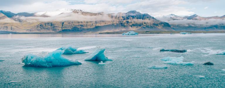 Icebergs are seen floating on a blue-green sea with mountains in the distance.