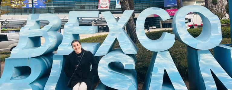 Paige Wise sits in front of a sign that says Bexco, Busan