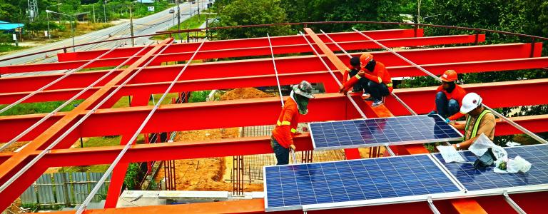 Workers install solar panels on top of red steel structure.