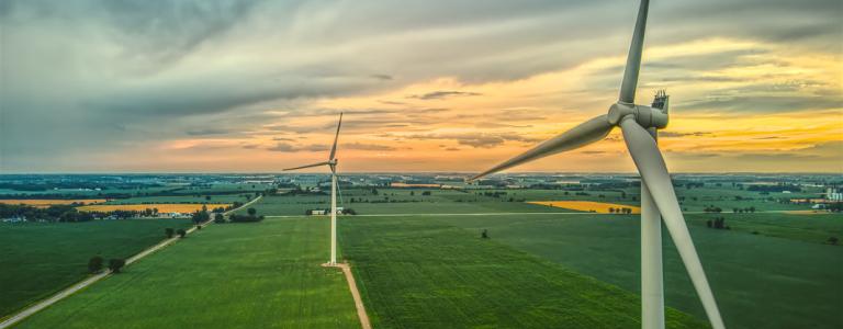 Wind turbines in a farm field. The sun sets in the horizon.
