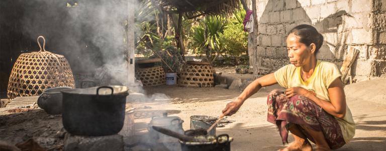 A woman crouches on the ground and stirs a pot while cooking outside.