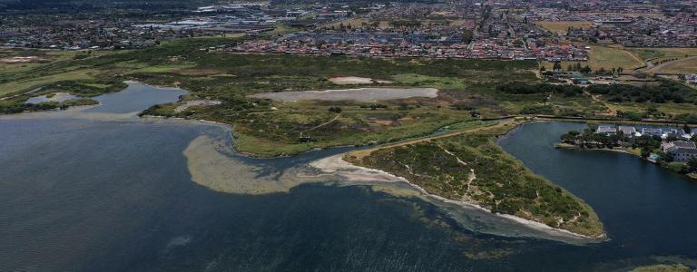 Water course next to sand and grass, urban area and mountain in the background.