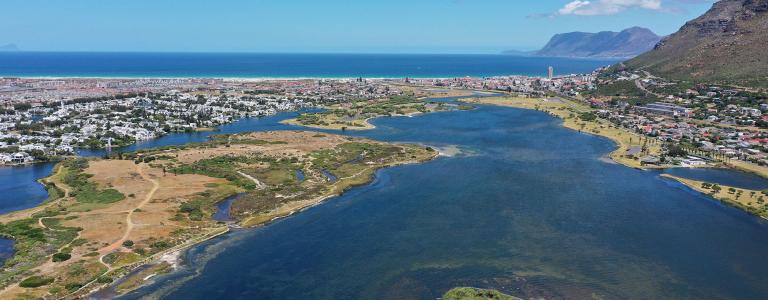 Water course next to sand and grass, urban area with buildings in the background.