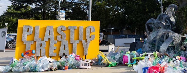 A yellow sign stands in a parking lot. It is surrounded by different items made of plastic in an art display. Text on the sign reads "Plastic Treaty."