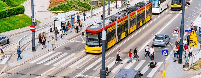 A tram and cars drive down a city street in Europe. Many pedestrians walk on the sidewalks and cross the street.
