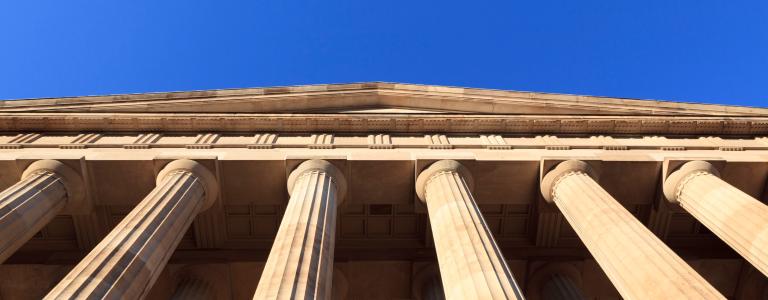 Low-angle view of a neoclassical building facade with tall fluted columns supporting an ornate entablature, photographed against a clear blue sky.