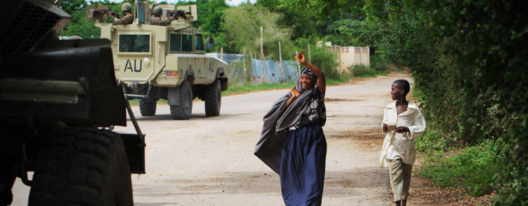 A woman and a young person walk along a dirt road, passing an armored military vehicle marked with "AU." Credit: AU-UN IST PHOTO / Stuart Price