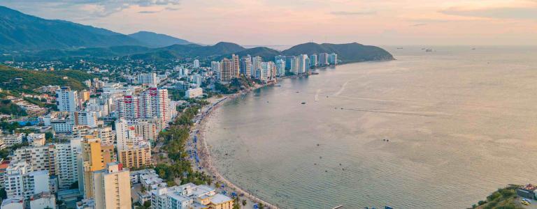 Aerial view of a coastal city with high buildings and a boulevard alongside a turquoise bay, as well as mountains in the background
