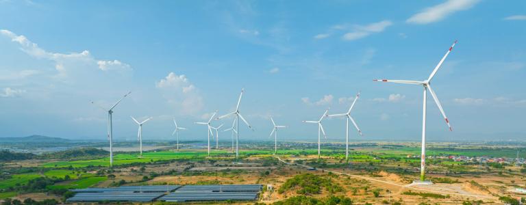 Aerial view of a wind and solar farm across a mixed landscape of dry scrubland and green agricultural fields, with a row of white wind turbines and a large solar panel array under a partly cloudy blue sky.