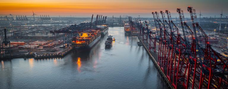 Aerial view of a container terminal with large container cargo ships and gantry cranes. A cargo ship is navigating through the central waterway. On both sides of the waterway are massive container terminals lined with red and black gantry cranes used for loading and unloading ships. on the left docked ships with brightly lit containers.