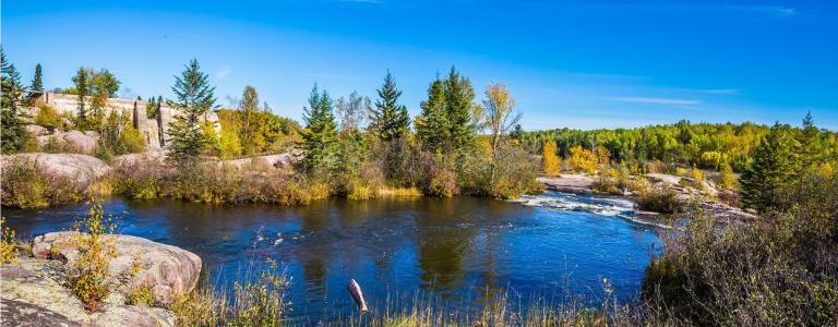 A basin of the Winnipeg River. There are a variety of large rocks, shrubs, and trees along the bank.