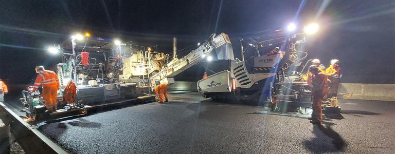 Construction workers laying asphalt at night with bright lights.