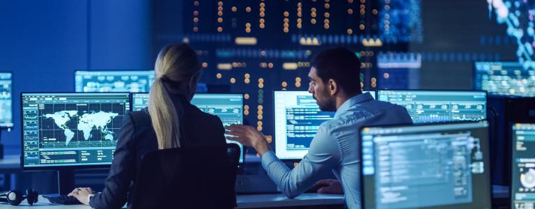 A man and woman sit at their computers in a room full of computers with data, comparing results.