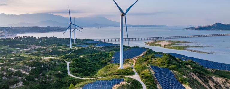 Aerial view of three wind turbines standing tall. Large arrays of solar panels occupy the fields below. There are mountains, water, and a bridge in the distance.