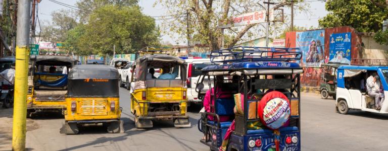 Several auto rickshaws and e-rickshaws with passengers are visible on a busy main street in Mathura, with commercial signage in Hindi, trees, and a wall mural in the background under daylight.