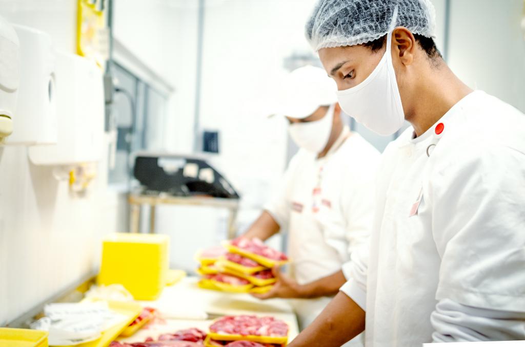 A man wearing a hairnet and mask handles raw meat in a brightly lit facility
