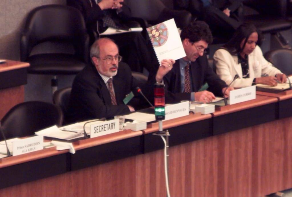 David Runnalls sits at a long conference table at a UN meeting holding up a book