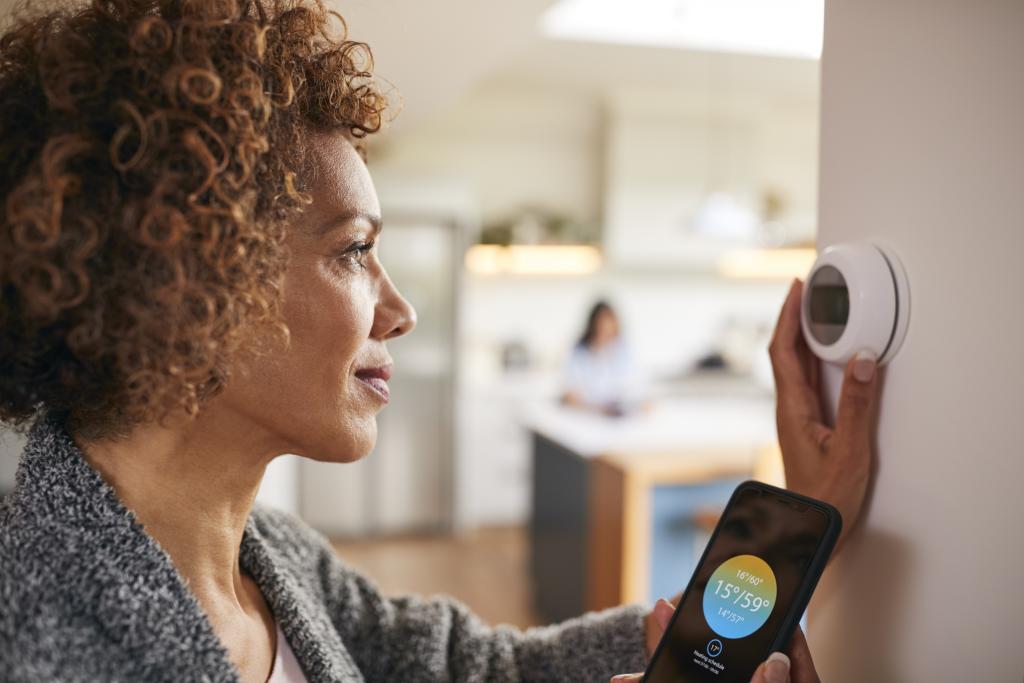 A Black middle-aged woman turns the dial on a smart thermostat while the temperature shows on her smart phone