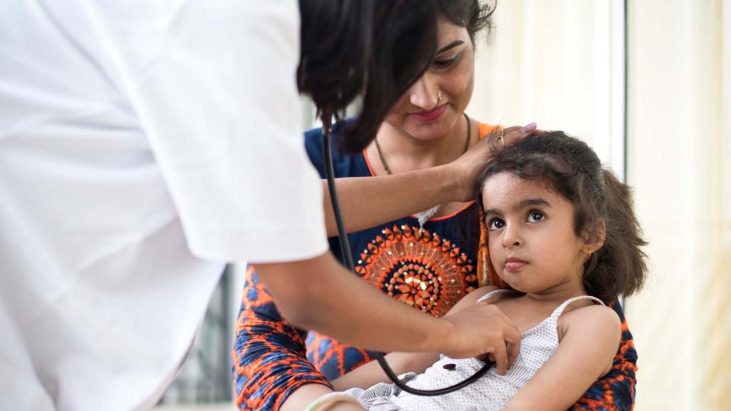 A woman in a health facility in India holds her daughter in her arms as a doctor undertakes a check-up.