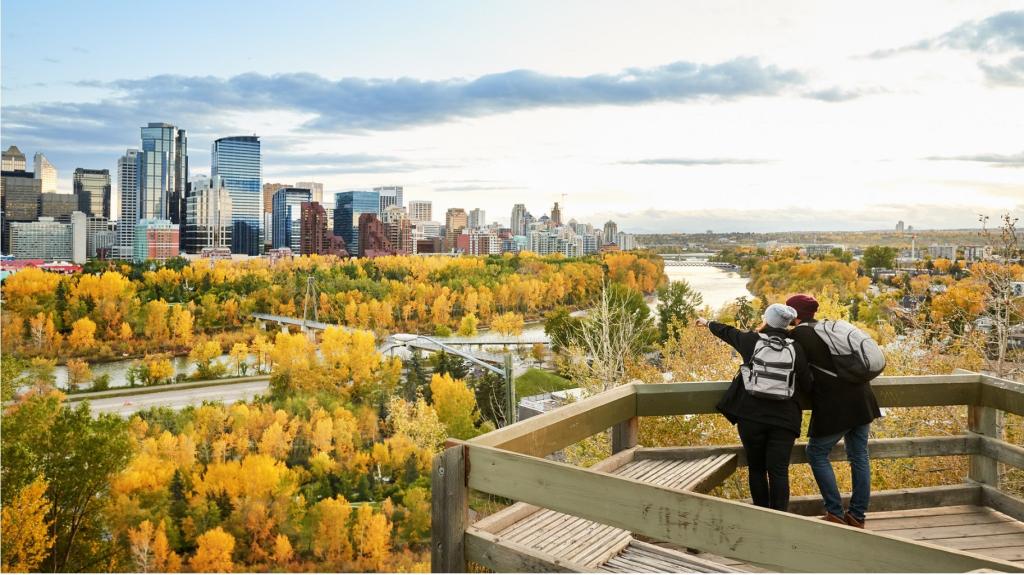 Two people stand on a wooden platform lookout point and look out into the distance. The platform overlooks a park during autumn with trees and a river running through it. There are city skyscrapers in the distance.