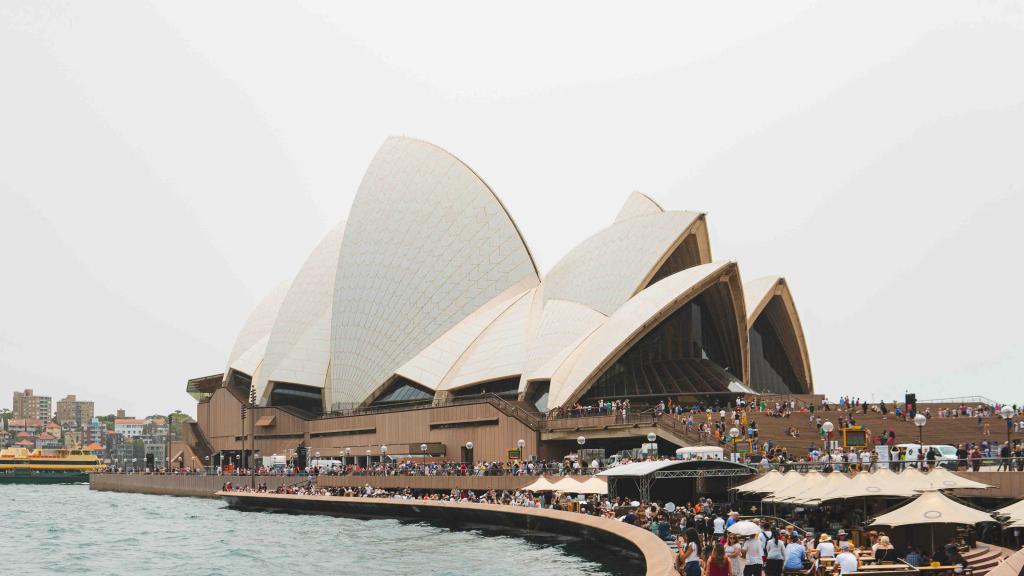 A crowd of people walk by the Sydney Opera House.