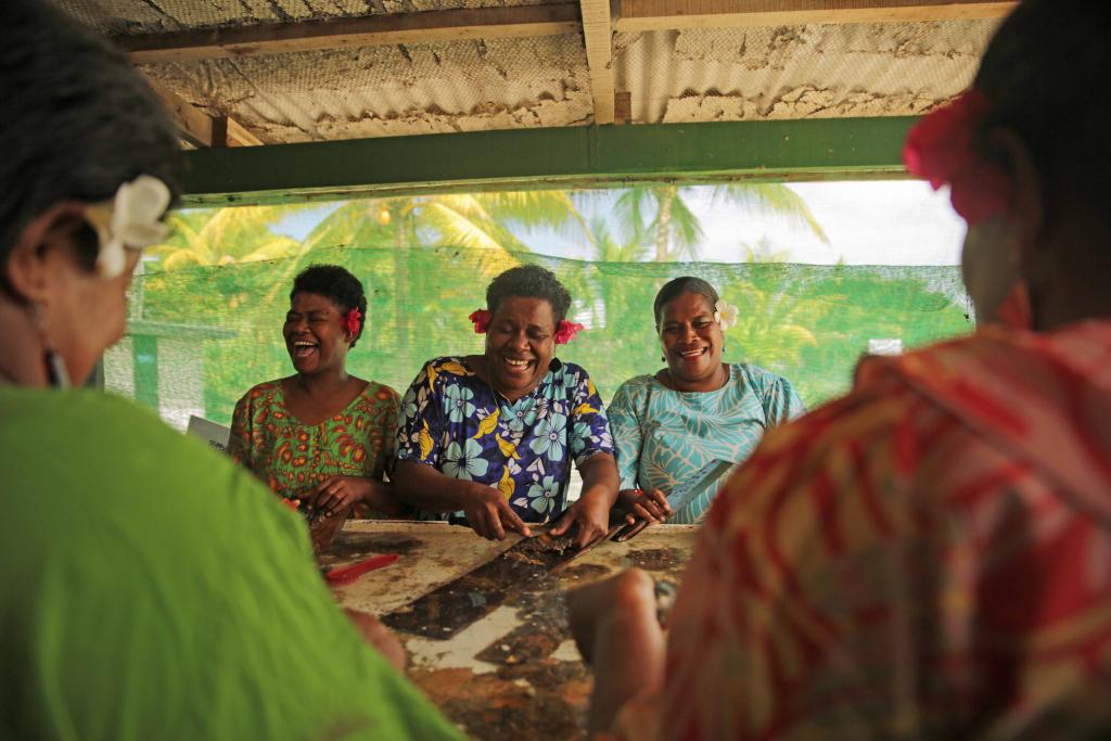 Three local women in Fiji laugh as they prepare oysters indoors. 