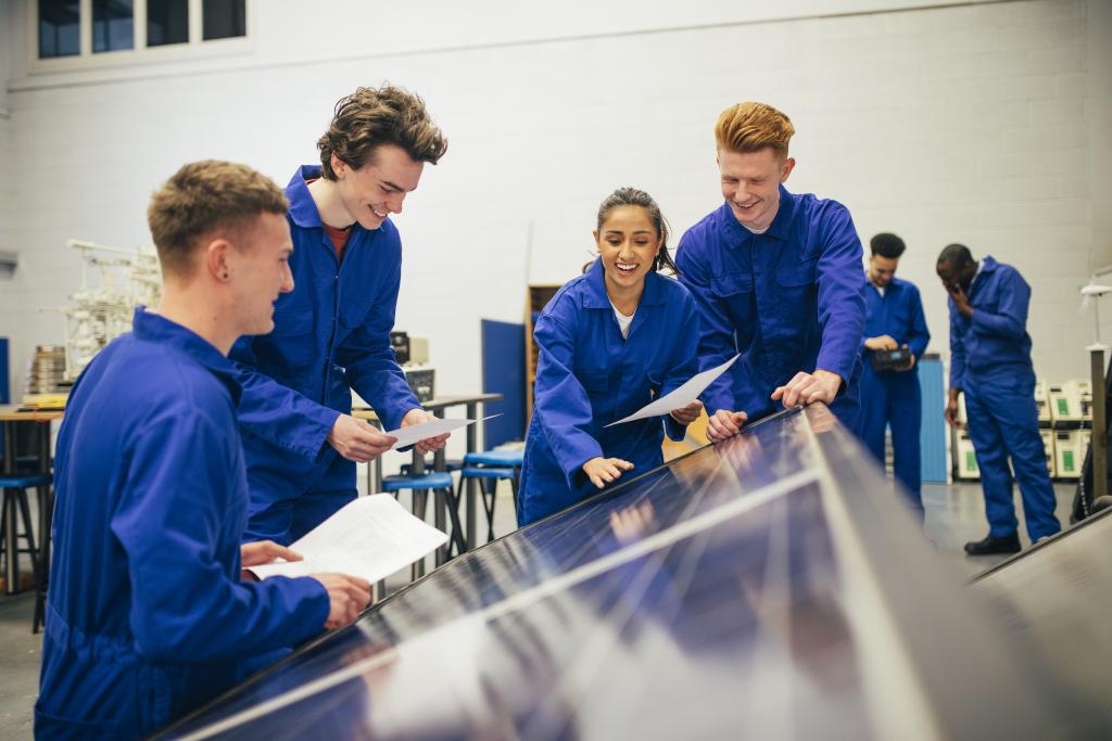 Four people in blue jumpsuits hold paper and smile while looking at a solar panel.