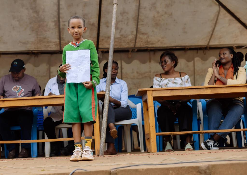 A young student stands on a stage holding a piece of paper and looks into the camera.