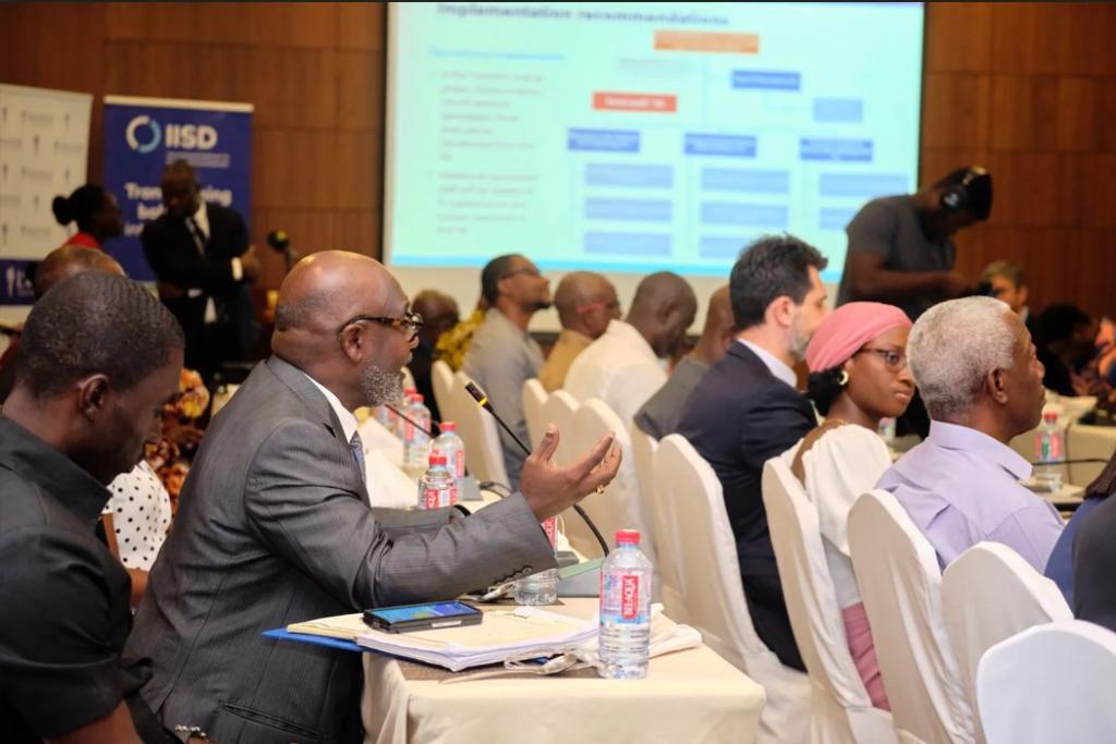 A meeting room with someone speaking in the foreground and a Powerpoint presentation in the background. Audience is listening.