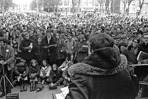 A black and white photo of the first Earth Day showing hundreds of people gathered outside to watch a man speaking