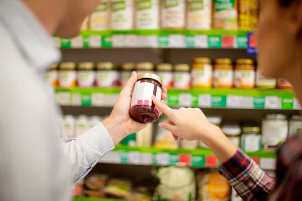 Close-up of a man holding a small glass jar in a grocery store. A woman beside him looks and points at something on the label.