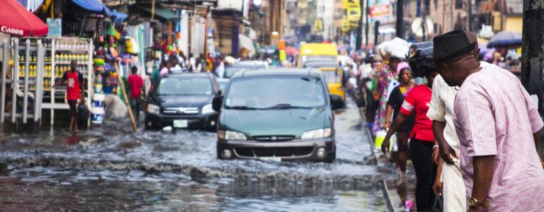 Cars drive down flooded market street in Lagos, Nigeria, with people crowded on either side.