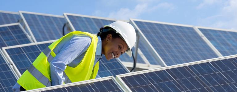 Female energy engineer works on solar panels in yellow vest and white helmet.