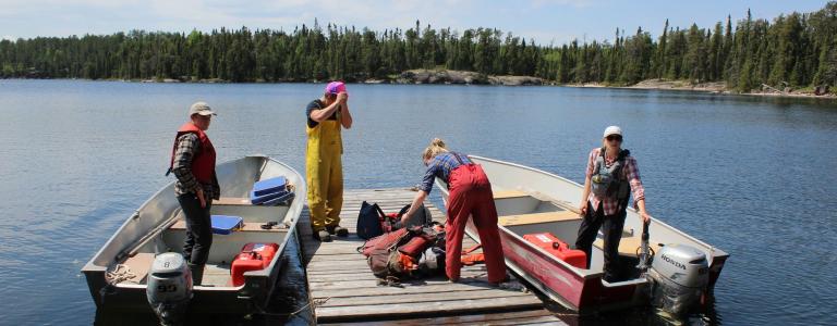 Four scientists stand in two boats floating on a lake on a sunny day