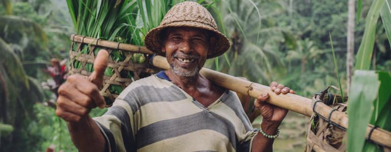 A middle-aged farmer wearing a hat holds a pole with a basket full of greenery while smiling at the camera