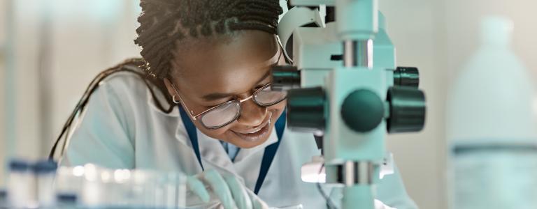 A Black woman in a lab coat looks through a microscope and smiles