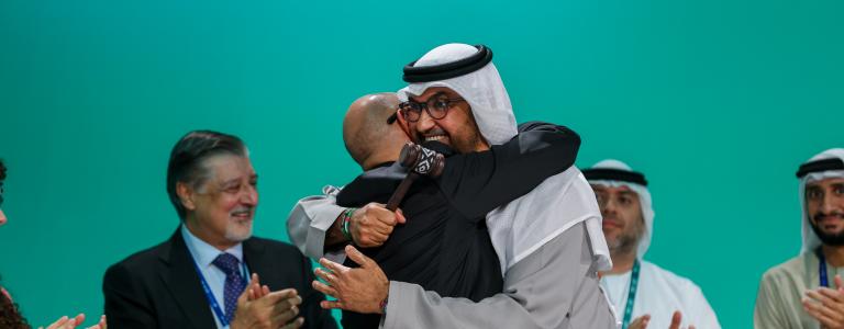 Two men, one in traditional Saudi dress and the other in a suit, embrace on the stage of COP 28 while others clap in the background
